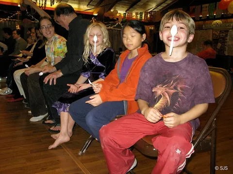 Children and adults seated in a row balancing spoons on their noses, laughing (Jesse, front)