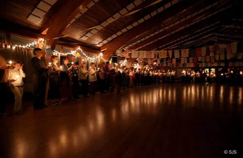 A long line of people holding lit candles in a dark hall decorated with colorful paper banners