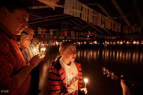A girl holding a lit candle among older adults also holding candles in a dim hall (Fiona)