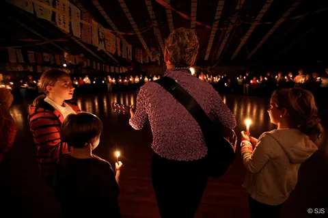 Three children and a woman with a guitar seen from behind, facing a large circle of people holding candles in a dim hall (Patricia Williams)