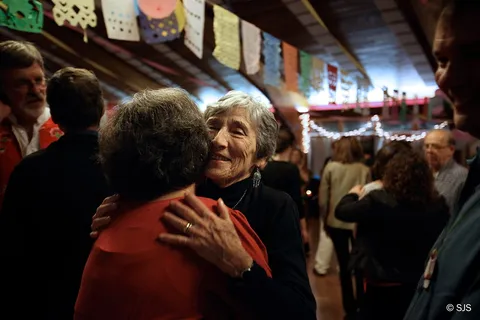Two older women embracing in a hug at an indoor gathering with paper banners overhead (Ruth Sherman and Unknown)