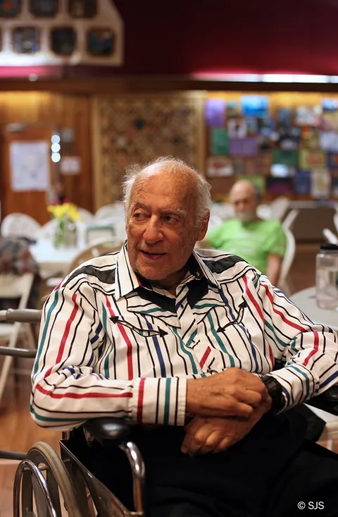 An elderly man in a striped shirt sits in a wheelchair, smiling, in a large hall (Alex Sherman)