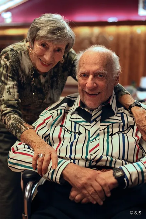 An older woman stands behind and embraces an elderly man in a wheelchair, both smiling (Ruth and Alex Sherman)