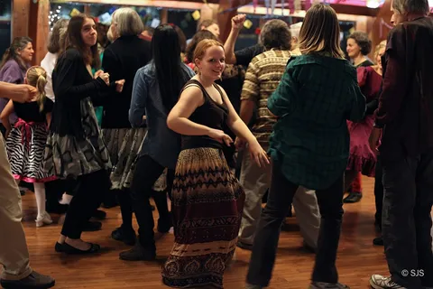 A person in a patterned skirt dances and smiles amid a crowded dance floor (Rain Woods)