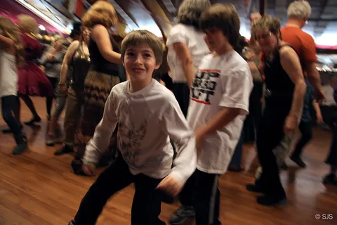 A boy dances energetically on a wooden dance floor among other children and adults (Jesse)