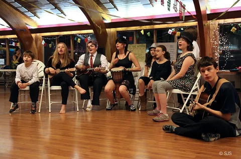 A group of children and teens sing and play ukuleles, guitar and a drum while seated in a row (From left to right: Jesse, Fiona, Crow, Momo, Unknown, Emily, Scout, and Max)