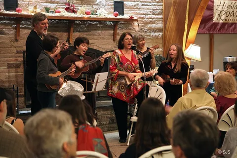 A group performs on ukuleles in front of a stone fireplace as an audience watches