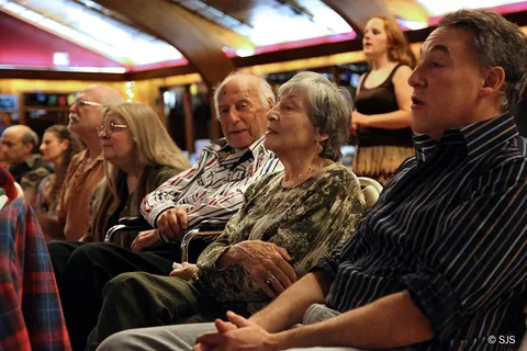 A row of seated people, some elderly, listen attentively in a large hall (From left to right: Alan and Glenn Lebwohl-Steiner, Alex, Ruth, and David Sherman, Rain Woods in the background)
