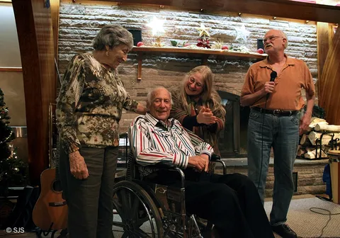 An elderly man in a wheelchair is surrounded by three smiling older adults near a decorated fireplace (From left to right: Ruth and Alex Sherman, Glenn and Alan Lebwohl-Steiner)
