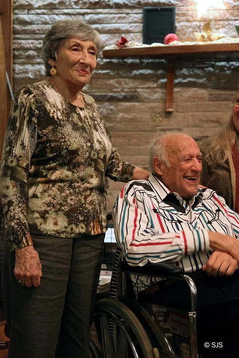 An older woman stands beside an elderly man seated in a wheelchair near a stone fireplace (Ruth and Alex Sherman, Glenn Lebwohl-Steiner almost out of frame)