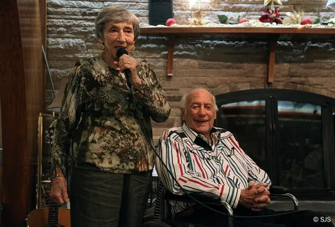 An older woman speaks into a microphone beside an elderly man seated in a wheelchair (Ruth and Alex Sherman)