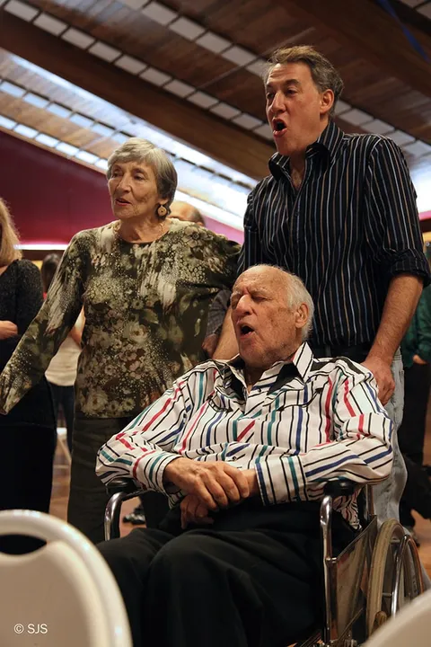 An elderly man in a wheelchair sings with a woman and man standing beside him (Ruth, Alex, and David Sherman)