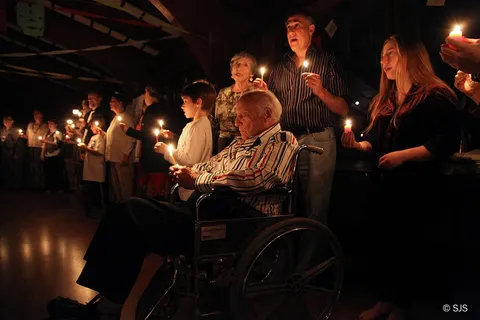 People hold lit candles in a darkened hall, an elderly man in a wheelchair in front (From left to right in foreground: Jesse, Ruth, Alex, and David Sherman, Fiona)