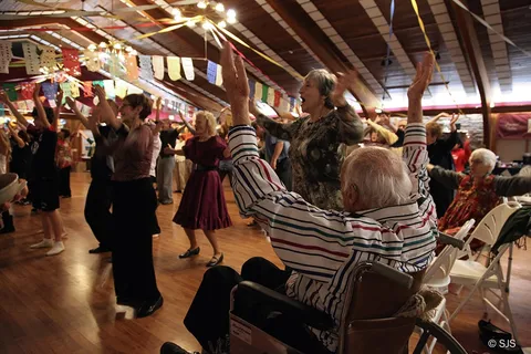 A crowd of dancers, including an elderly man in a wheelchair, raise their arms together dancing YMCA (Ruth and Alex Sherman)