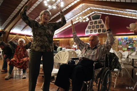 A woman and an elderly man in a wheelchair raise their arms during a group dance (Ruth and Alex Sherman)