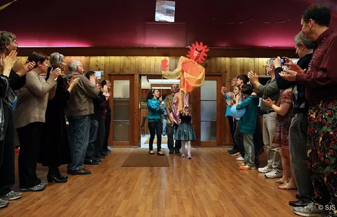 A crowd claps as two children and a man carry a tall orange sun-faced puppet on poles through a doorway