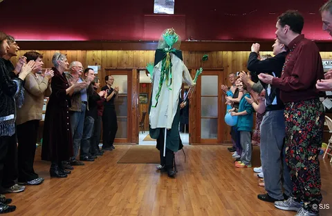 A crowd applauds as a tall green-robed puppet figure on stilts walks through the hall