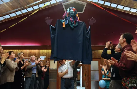 A crowd applauds as a person carries a large draped puppet head with outstretched hands overhead