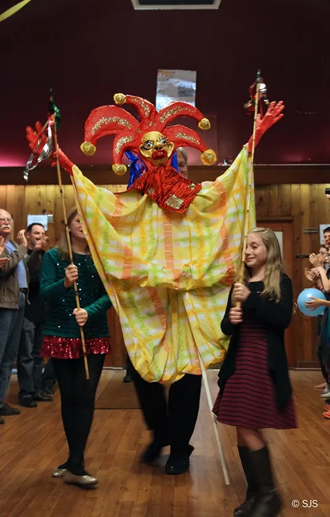 A tall jester puppet in a red and gold costume is carried through a crowd by two girls holding poles