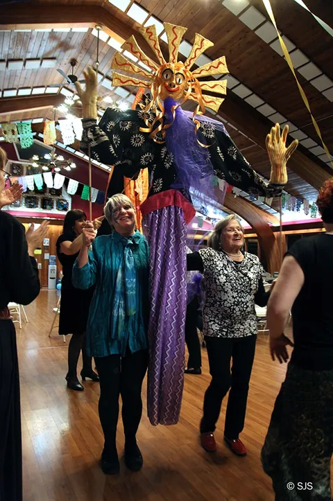 A tall sun-faced puppet on stilts in purple robes stands between two smiling women holding its poles