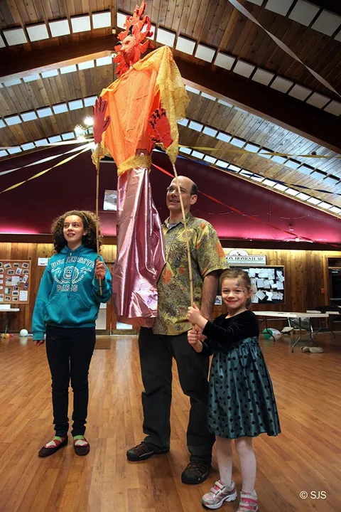 A man and two girls pose with a tall orange sun puppet on a pole indoors