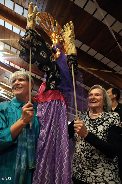 Two smiling women hold poles supporting a puppet with golden hands and a purple and black robe (Kay and Jane Gering)