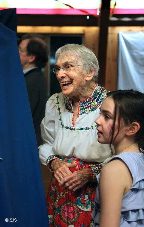 An elderly woman in an embroidered blouse laughs beside a young girl with painted face (Helen Hardin)