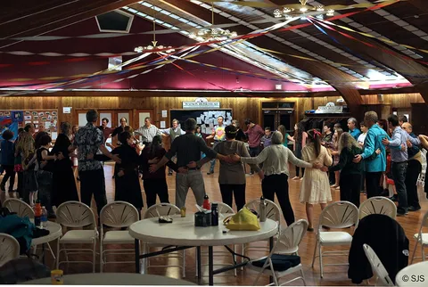 Large group dancing in a circle holding raised arms in a decorated wooden hall