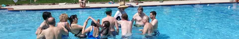 Group of people playing in a circle in a swimming pool, some shirtless, on a sunny day