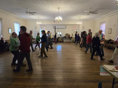 Two parallel lines of people dancing across a wooden-floored hall with ceiling fans and chandeliers