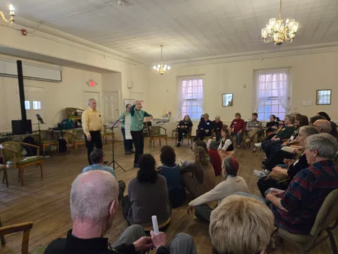 Two people teaching a song to a seated circle of people in a community hall