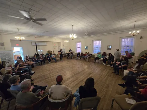 A man playing guitar and singing to a seated circle of people in a large hall