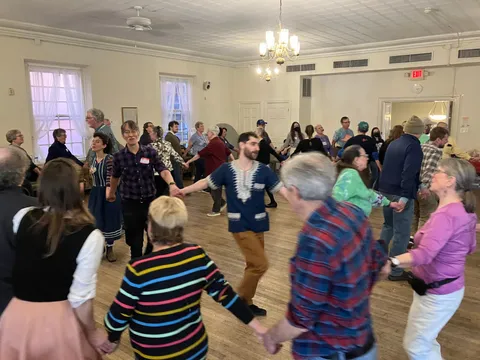 A large circle of people holding hands mid-dance in a hall with white walls and chandeliers