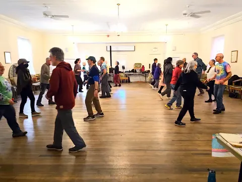 People walking in a line dance across a wooden floor past chairs and tables