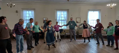A circle of dancers holding hands in a bright hall with tall curtained windows